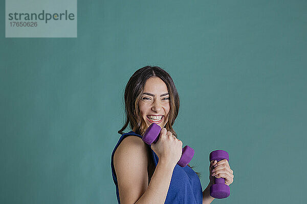 Cheerful woman with purple dumbbells standing against blue background