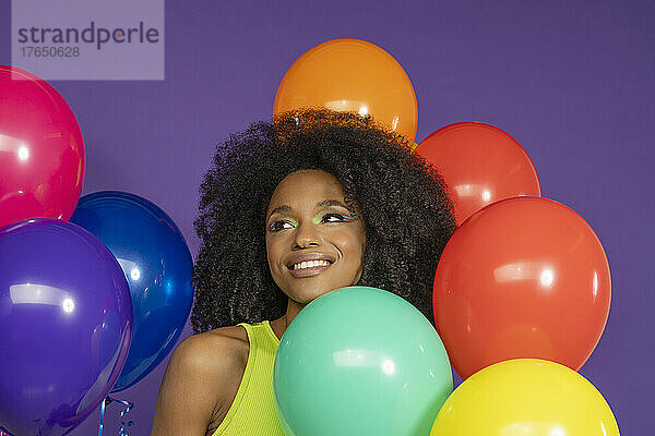 Lächelnde junge Frau mit lockigem Haar inmitten bunter Luftballons vor violettem Hintergrund