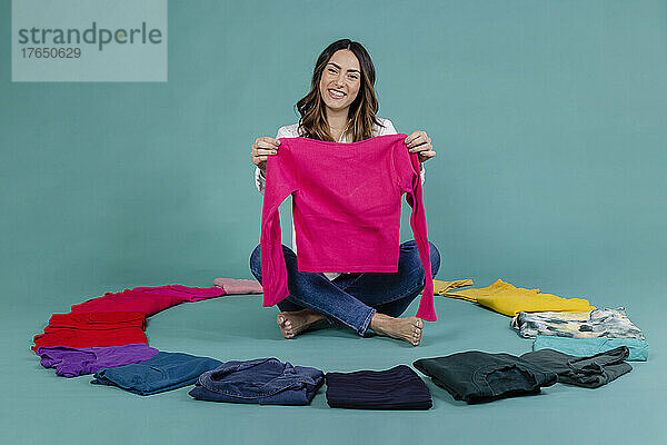 Smiling woman holding pink t-shirt sitting cross-legged amidst clothes against blue background