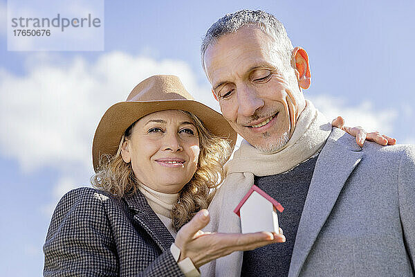 Lächelndes reifes Paar hält an einem sonnigen Tag ein kleines Hausmodell in der Hand