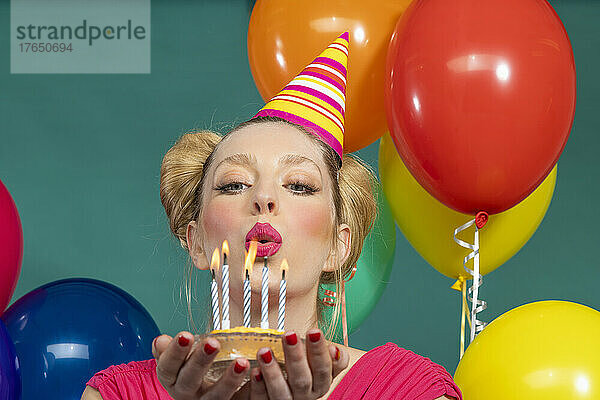 Schöne junge Frau mit Partyhut bläst Kerzen auf Geburtstagstorte vor grünem Hintergrund