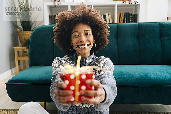 Glückliche junge Frau  die zu Hause am Sofa sitzt und Weihnachtsgeschenk überreicht