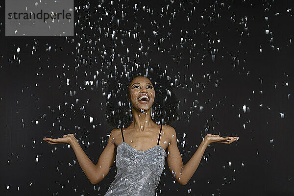 Happy young woman with mouth open standing under falling confetti against black background