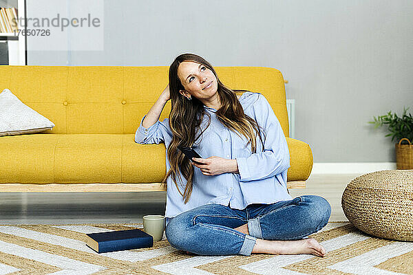Nachdenkliche Frau mit Mobiltelefon sitzt zu Hause auf dem Teppich neben dem Sofa
