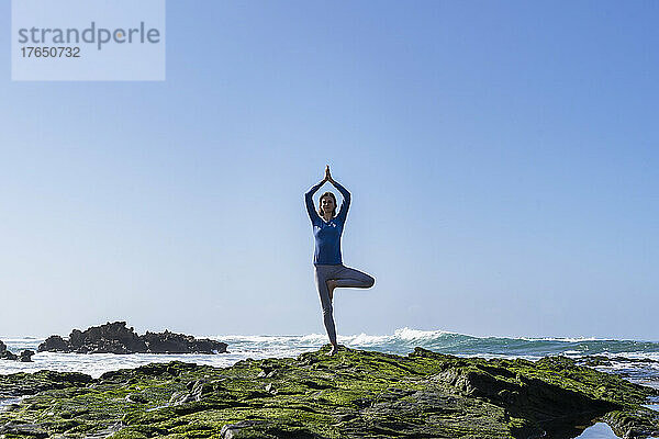 Frau praktiziert Baumpose-Yoga auf einem Felsen am Strand