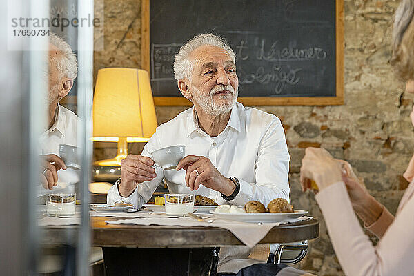 Senior man holding coffee cup sitting with woman at breakfast table in boutique hotel