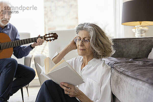 Ältere Frau mit der Hand im Haar liest Buch von einem Mann  der zu Hause Gitarre spielt