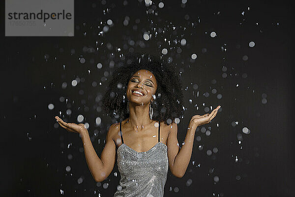 Smiling young woman with eyes closed standing under falling confetti against black background