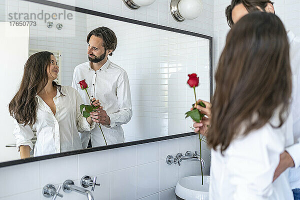 Man presenting woman with red rose in front of mirror