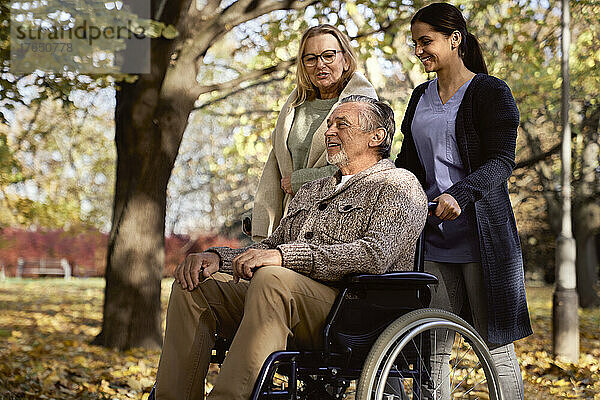 Lächelnde ältere Frau im Gespräch mit einem Mann  der im Rollstuhl sitzt und von einer Krankenschwester im Park geschoben wird