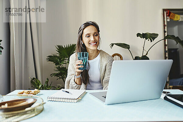 Glücklicher Freiberufler hält ein Glas Wasser in der Hand und arbeitet zu Hause am Laptop