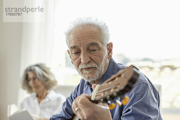 Älterer Mann spielt Gitarre mit Frau im Hintergrund zu Hause