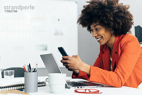 Glückliche Geschäftsfrau sitzt mit Laptop am Schreibtisch im Büro und nutzt ihr Smartphone