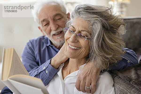 Älterer Mann umarmt Frau  die zu Hause im Wohnzimmer ein Buch liest