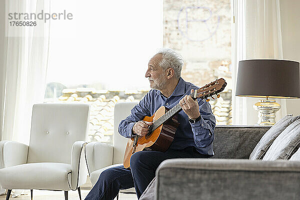 Älterer Mann spielt Gitarre und sitzt zu Hause auf dem Sofa im Wohnzimmer