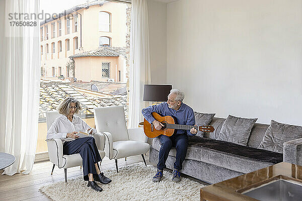 Älterer Mann spielt Gitarre und schaut Frau zu  die im Wohnzimmer ein Buch liest