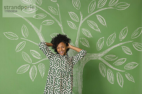Glückliche junge Frau mit der Hand im Haar steht vor einem Baum und zeichnet an einer grünen Wand