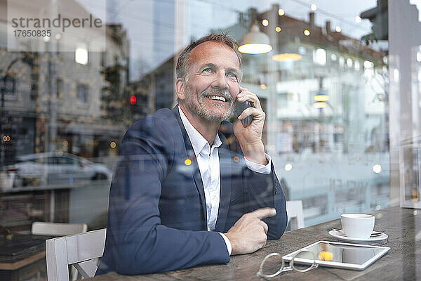 Lächelnder Geschäftsmann  der mit dem Handy spricht und durch das Fenster im Café blickt