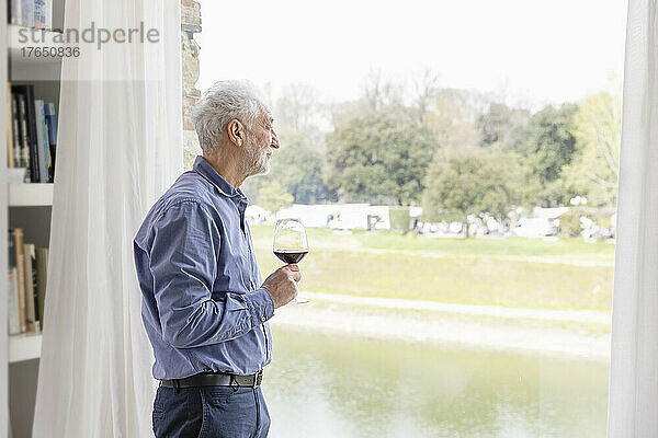Älterer Mann hält Weinglas in der Hand und schaut zu Hause durch das Fenster