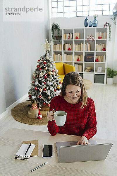 Glückliche Frau mit Laptop und Kaffeetasse sitzt zu Hause im Wohnzimmer