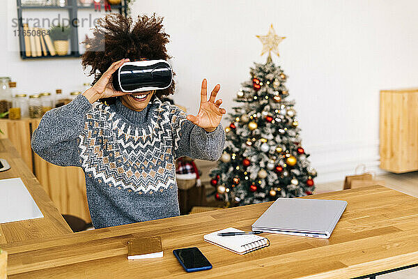 Lächelnde Frau mit Virtual-Reality-Simulator gestikuliert am Tisch im Wohnzimmer