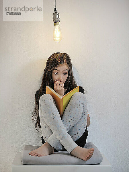 Neugieriges Mädchen liest ein Buch und sitzt unter einer Glühbirne vor der Wand