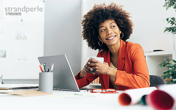 Lächelnde Geschäftsfrau mit Kaffeetasse und Laptop am Schreibtisch im Büro