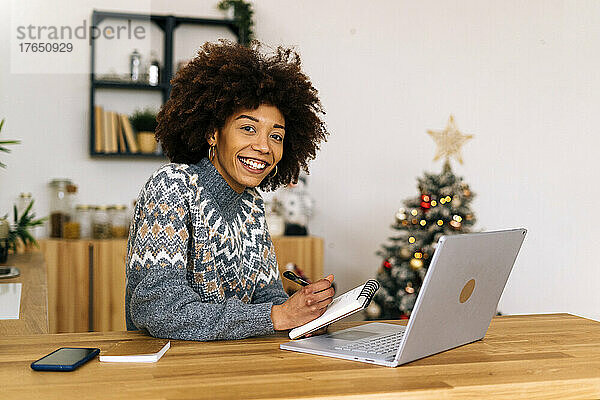 Glückliche junge Frau mit Notizblock und Laptop sitzt am Tisch im Wohnzimmer