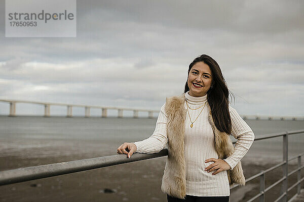 Lächelnde Frau mit der Hand auf der Hüfte  die am Geländer vor dem Meer steht