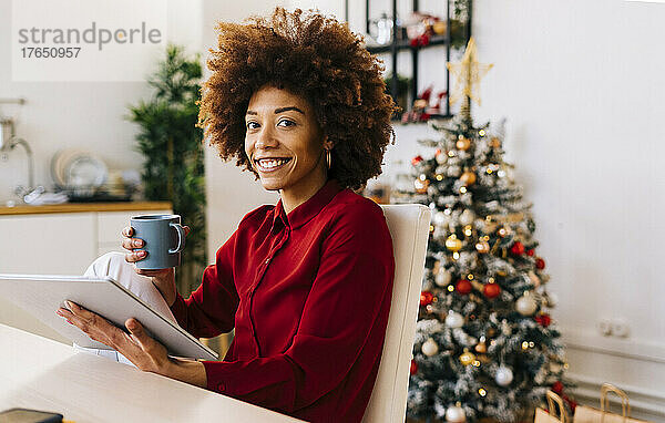 Glückliche Frau mit Tablet-PC und Kaffeetasse sitzt zu Hause im Wohnzimmer