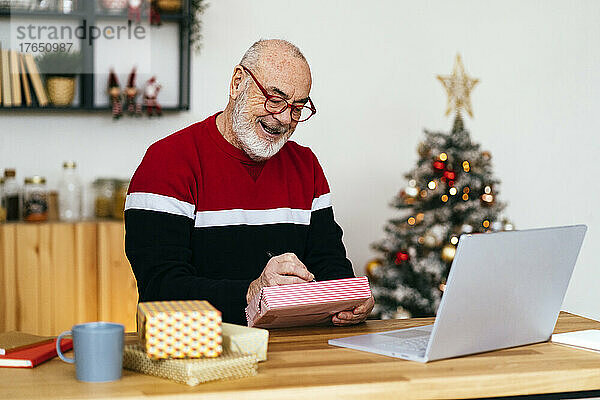 Glücklicher älterer Mann  der zu Hause mit Laptop am Tisch sitzt und auf eine Geschenkbox schreibt