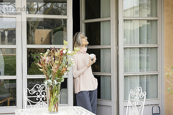 Nachdenkliche ältere Frau mit Kaffeetasse steht vor der Tür