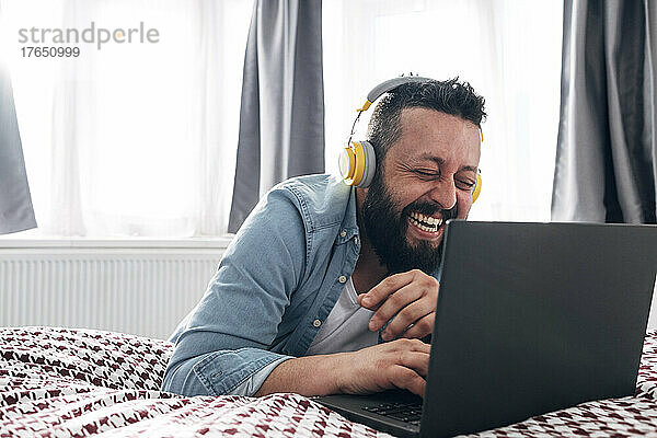Glücklicher Mann mit kabellosen Kopfhörern  der zu Hause einen Laptop benutzt