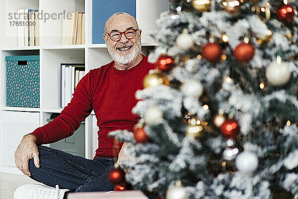 Lächelnder älterer Mann mit Brille blickt im Wohnzimmer auf den Weihnachtsbaum