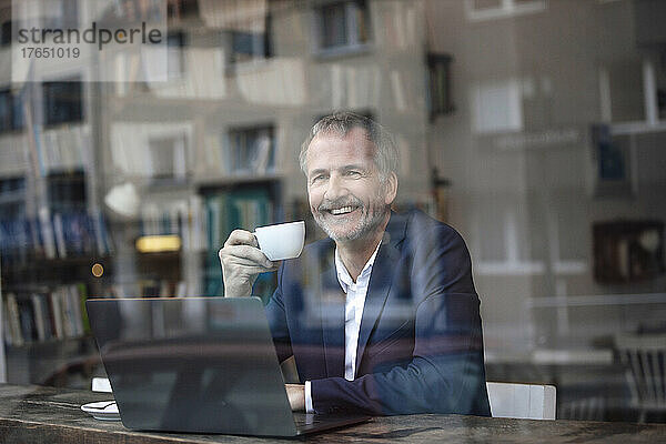 Glücklicher Freiberufler mit Kaffeetasse und Laptop  gesehen durch Glasfenster im Café