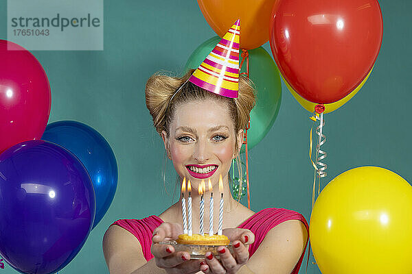 Fröhliche junge Frau mit Partyhut hält Geburtstagstorte inmitten von Luftballons vor grünem Hintergrund