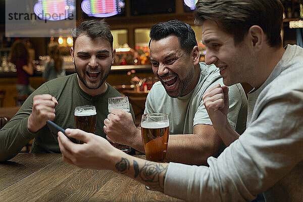 Drei fröhliche männliche Fußballfans trinken Bier und schauen sich ein Spiel auf dem Smartphone in einer Kneipe an