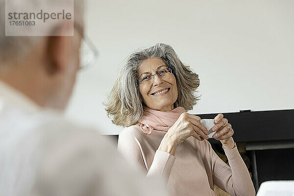 Lächelnde ältere Frau hält Kaffeetasse in der Hand und blickt Mann im Wohnzimmer an