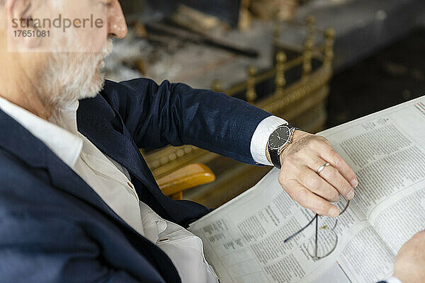 Älterer Mann mit Zeitung  der im Wohnzimmer sitzt und die Uhrzeit überprüft