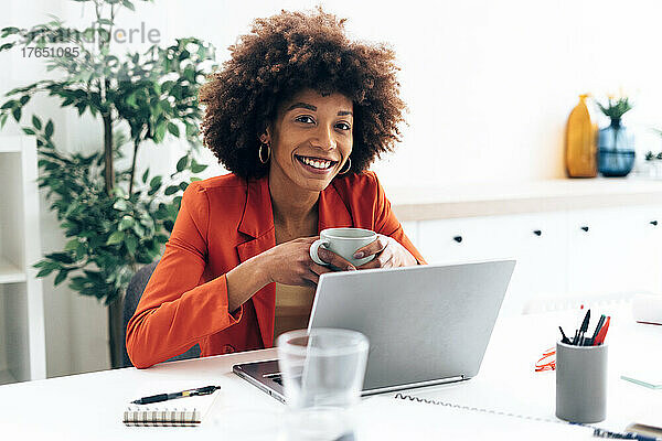Lächelnde Geschäftsfrau mit Kaffeetasse und Laptop am Schreibtisch