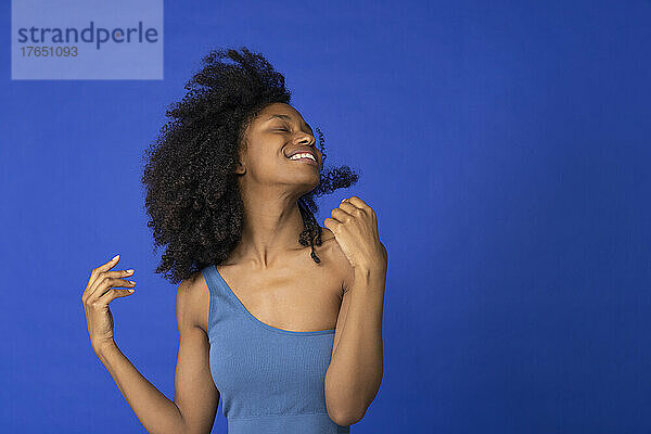 Fröhliche junge Frau mit geschlossenen Augen tanzt vor blauem Hintergrund