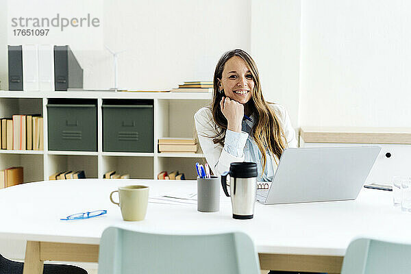 Lächelnde Geschäftsfrau mit Laptop sitzt mit der Hand am Kinn am Schreibtisch im Büro