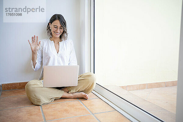 Glücklicher Freiberufler winkt bei einem Videoanruf über den Laptop und sitzt mit gekreuzten Beinen am Fenster