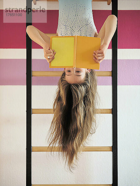 Girl reading book hanging upside down on wall bars