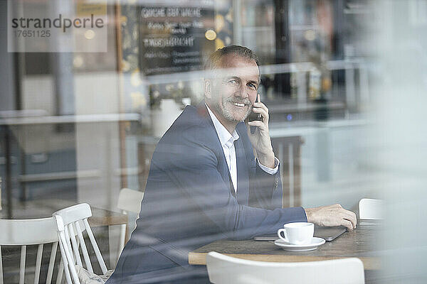 Lächelnder Geschäftsmann  der mit dem Mobiltelefon spricht und durch die Glasscheibe im Café gesehen wird
