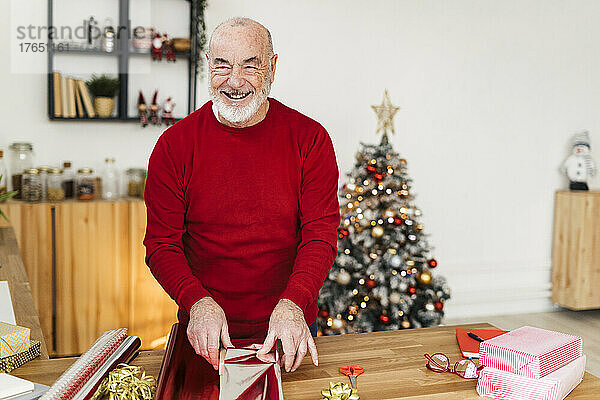 Cheerful senior man wrapping Christmas presents on table at home