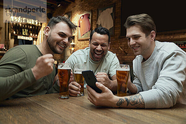 Drei fröhliche männliche Fußballfans trinken Bier und schauen sich ein Spiel auf dem Smartphone in einer Kneipe an