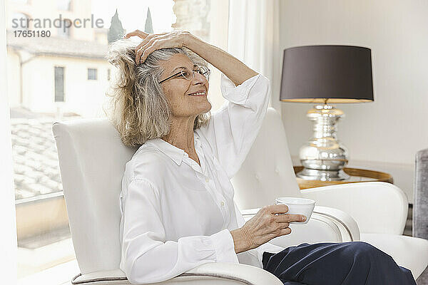 Lächelnde Frau mit der Hand im Haar und einer Kaffeetasse  die zu Hause auf einem Sessel sitzt
