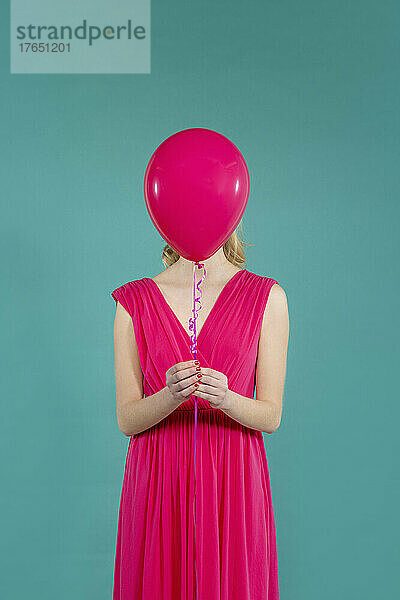 Junge Frau hält einen rosa Luftballon vor dem Gesicht und steht vor grünem Hintergrund
