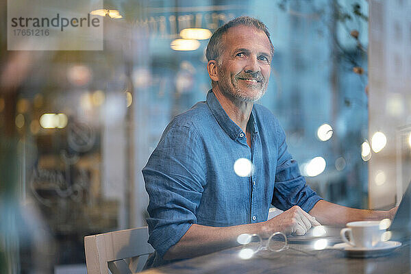 Lächelnder  nachdenklicher Geschäftsmann mit Laptop  der am Tisch im Café sitzt